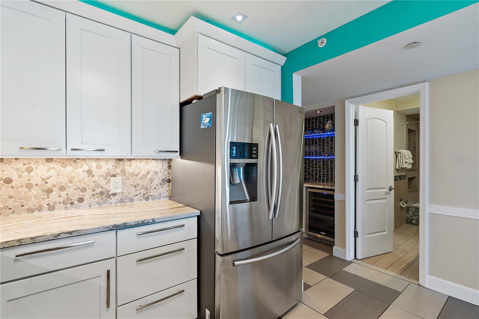 There is a closet pantry located to the left of the dry bar, out of sight. The open door leads to the second bathroom. Other features of the kitchen include a French door refrigerator, stone backsplash, and raised ceiling.