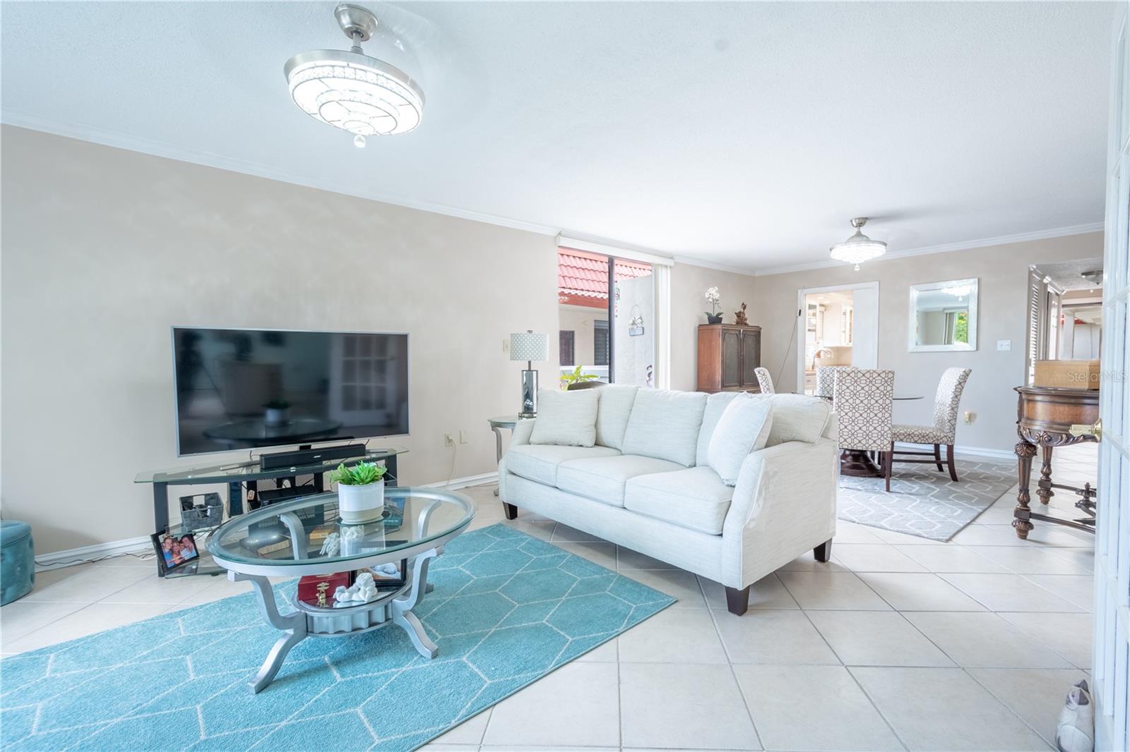 The living room is bathed in neutral tones. It features crown molding, ceramic tile flooring and and a stylish crystal ceiling fan with a light kit.