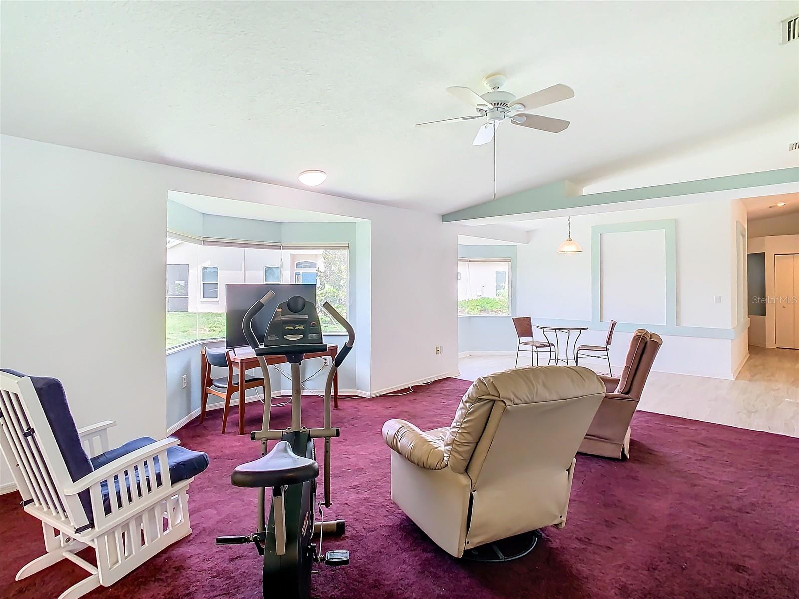 Living room sits on carpet and has a ceiling fan. Lots of natural light from the bay window.