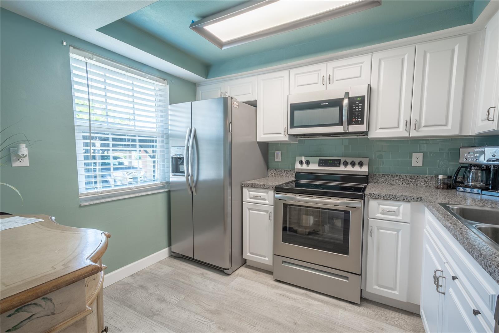 The kitchen features neutral-tone wood laminate flooring and a full suite of stainless steel appliances, including a dishwasher, range, microwave, and French door refrigerator with water and ice dispenser. There is room for a small breakfast table and a kitchen window brings in plenty of natural light.