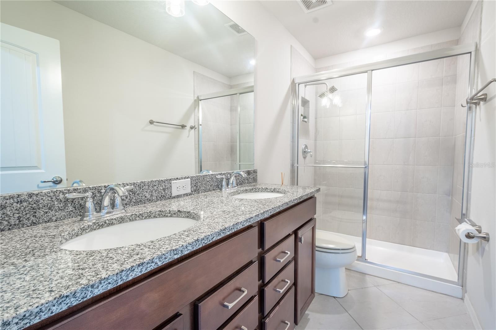 Bathroom 2 features a mirrored wood vanity with storage, dual sinks, granite countertop, ceramic tile floor and walk-in shower.