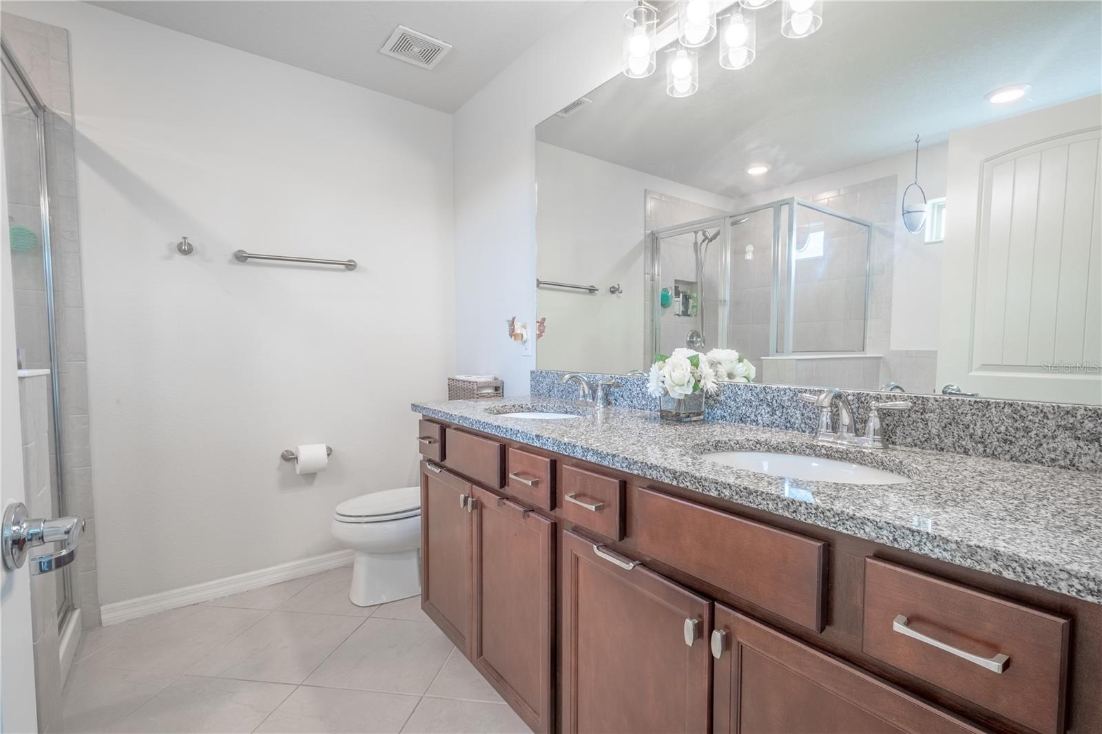 The primary bath features a mirrored wood vanity with storage, a downlight fixture, dual sinks, granite countertop, ceramic tile floor.