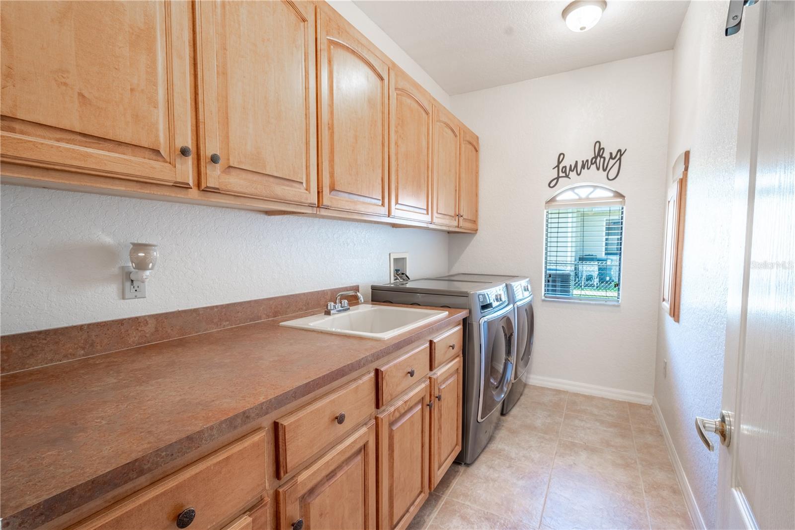 The laundry room features wood cabinets, a porcelain sink, plenty of room on the counter for folding, a ceramic tile floor and a stainless steel washer and dryer.