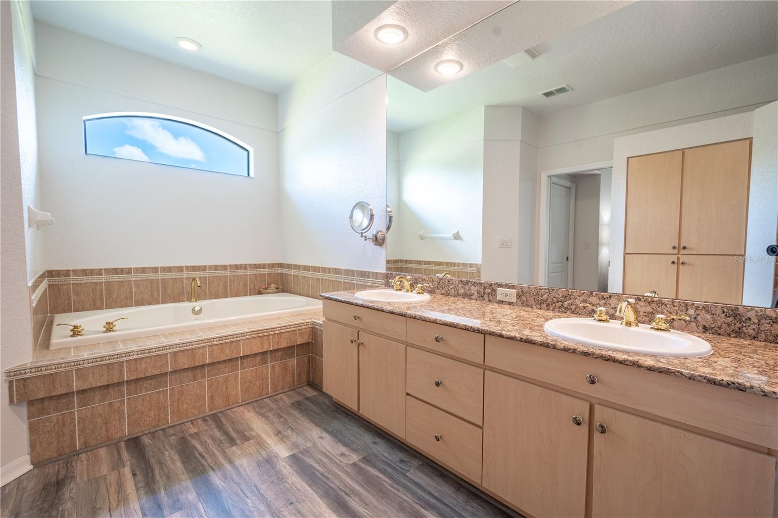 The primary ensuite bath features wood laminate flooring, a mirrored vanity with dual sinks, and a garden tub with transom window above.