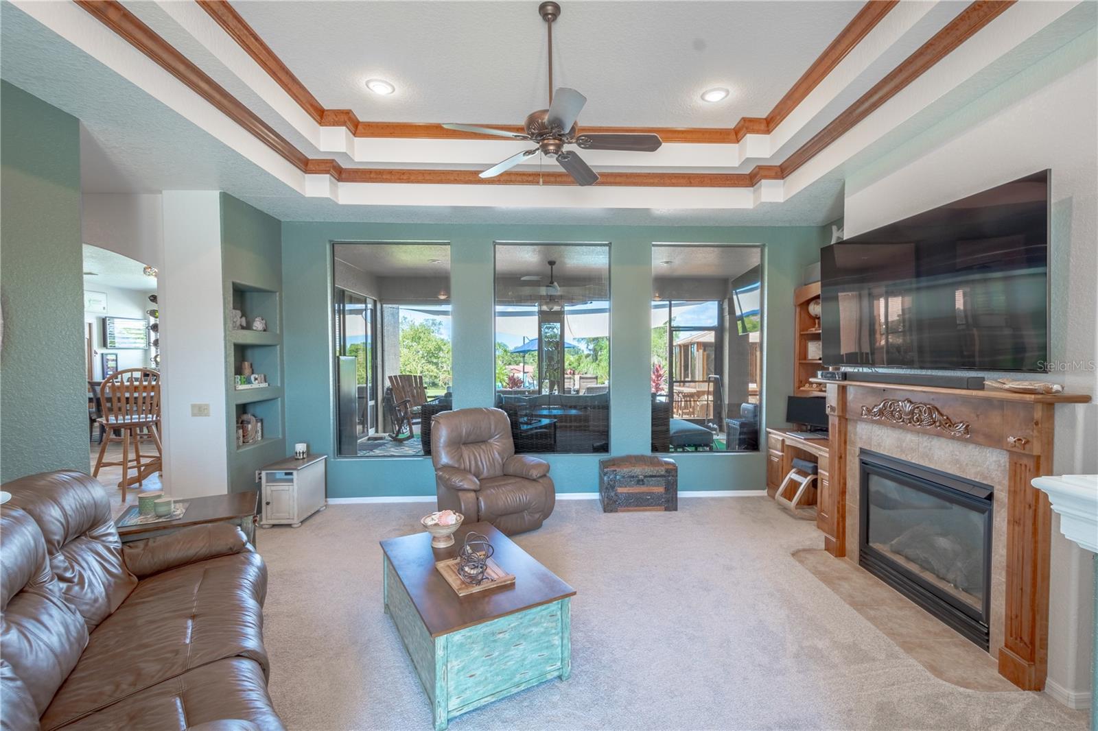 The living room features a double tray ceiling with rich wood molding, a ceiling fan, built in shelves and three floor to ceiling windows.