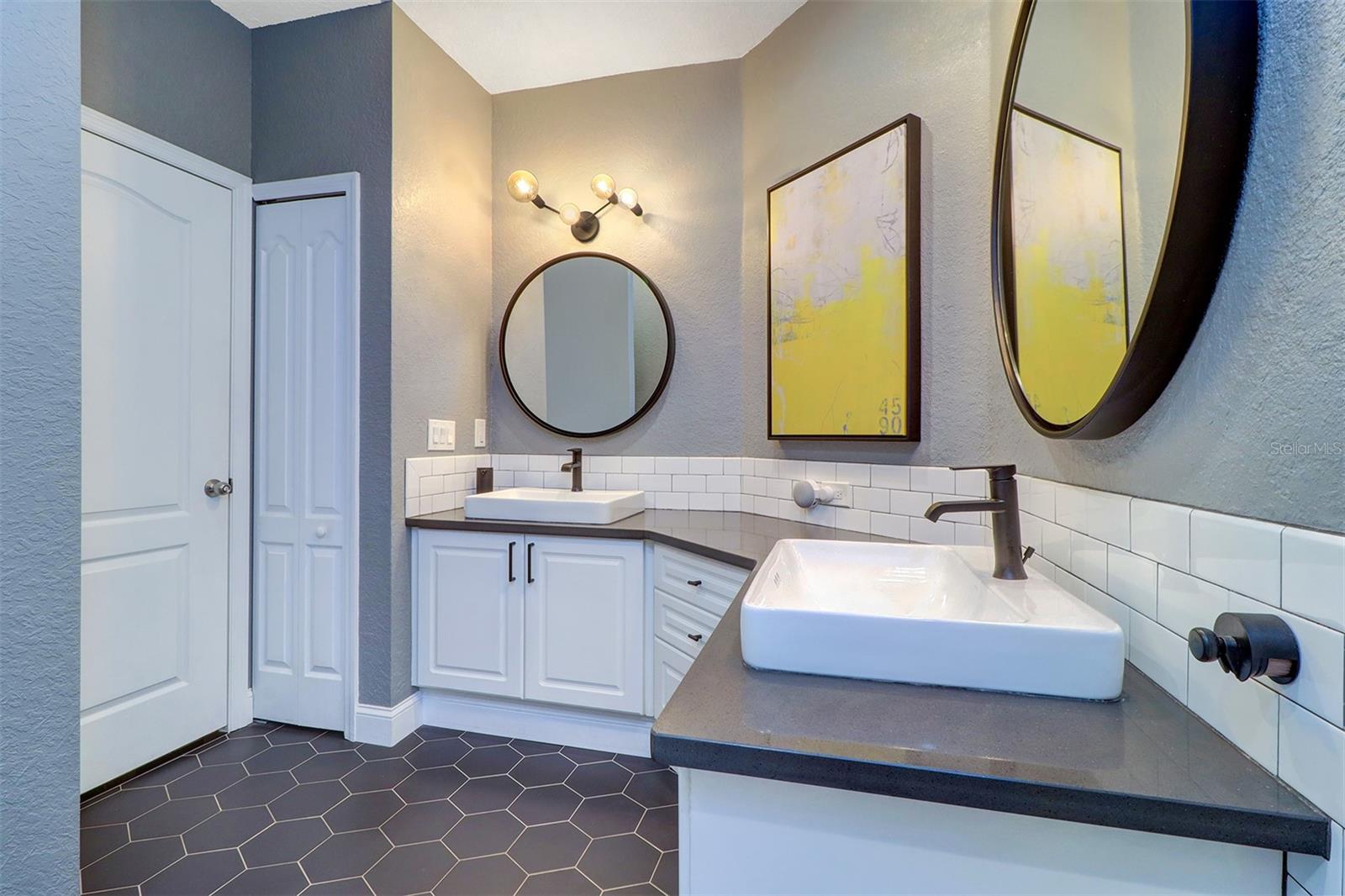 Large angled double vanity in primary bath with white subway tile backspalsh, graphite ceramic hexagon floor, round mirrors, black matte hardware and bathroom fixtures