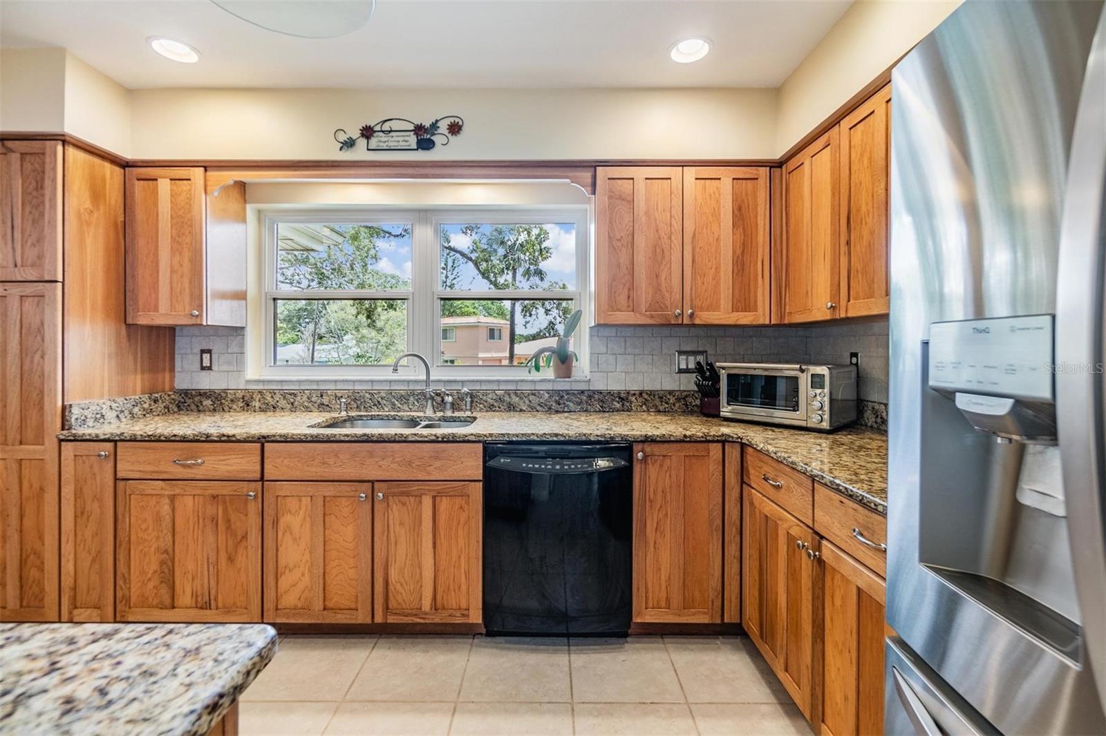 Kitchen with ample storage and counter space, granite countertops, gas stove, stainless steel refrigerator, and double windows overlooking front landscaping and circular driveway.