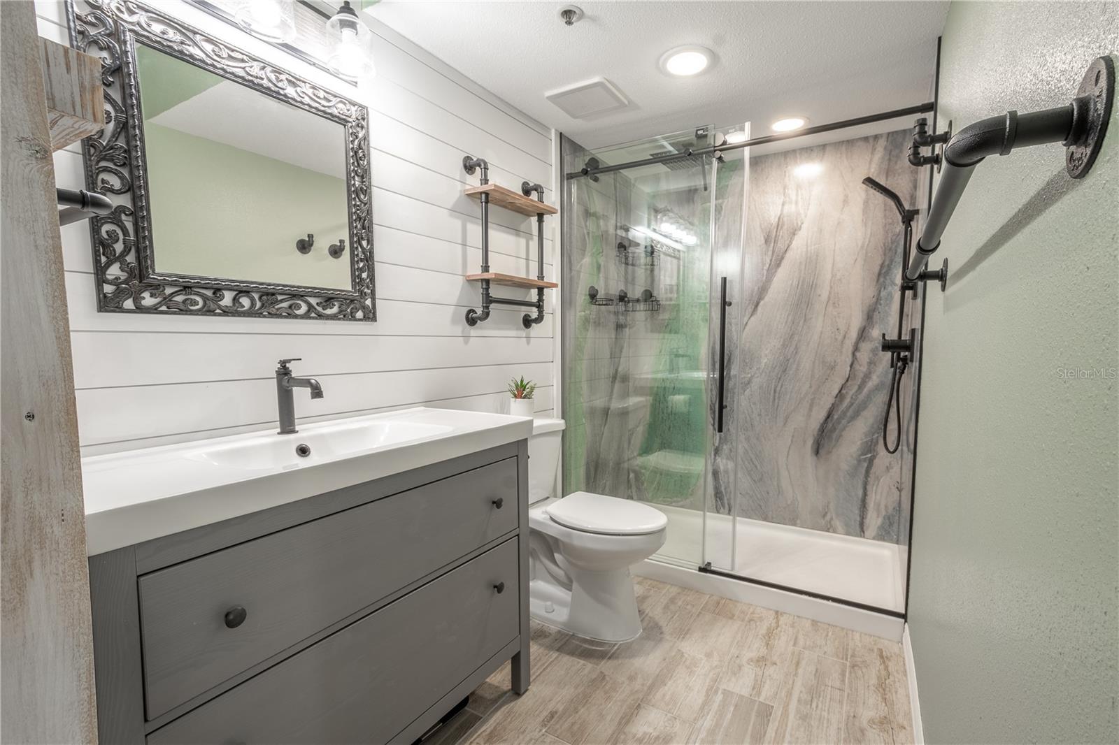 The primary ensuite bathroom features a mirrored vanity with storage and downlight fixture, a wood plank accent wall, wood plank tile flooring, and a shower with glass partition.