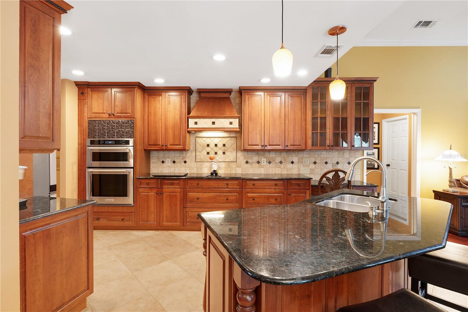 Kitchen features in wall Kitchenaid Dual ovens, cooktop with custom hood, a wood paneled dishwasher and sink built into a stunning island topped with granite, cherry wood cabinets with several deep drawers, a glass display cabinet and well crafted moldings.