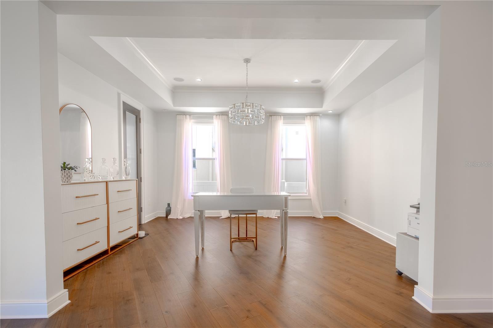 The dining room features a warm wood floor, neutral tones, and two windows adorned with elegant drapes, all illuminated by a striking chandelier.