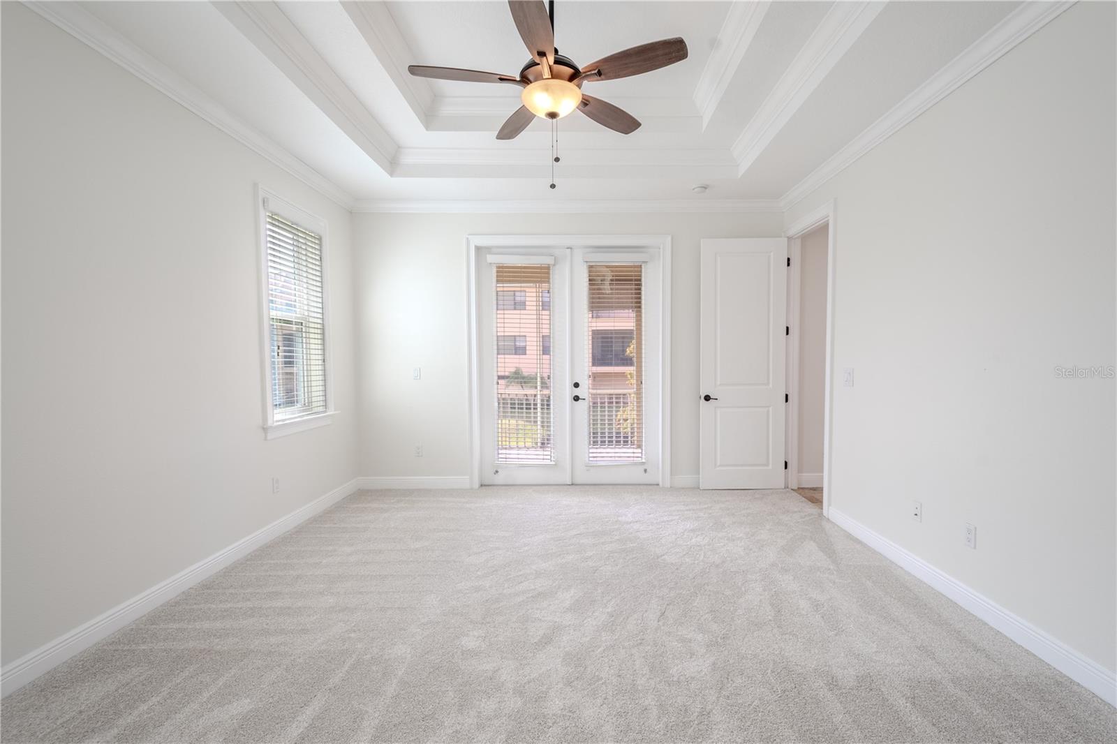 The Primary bedroom features new, neutral tone carpet, a tray ceiling with ceiling fan, crown molding and French doors leading to the balcony.