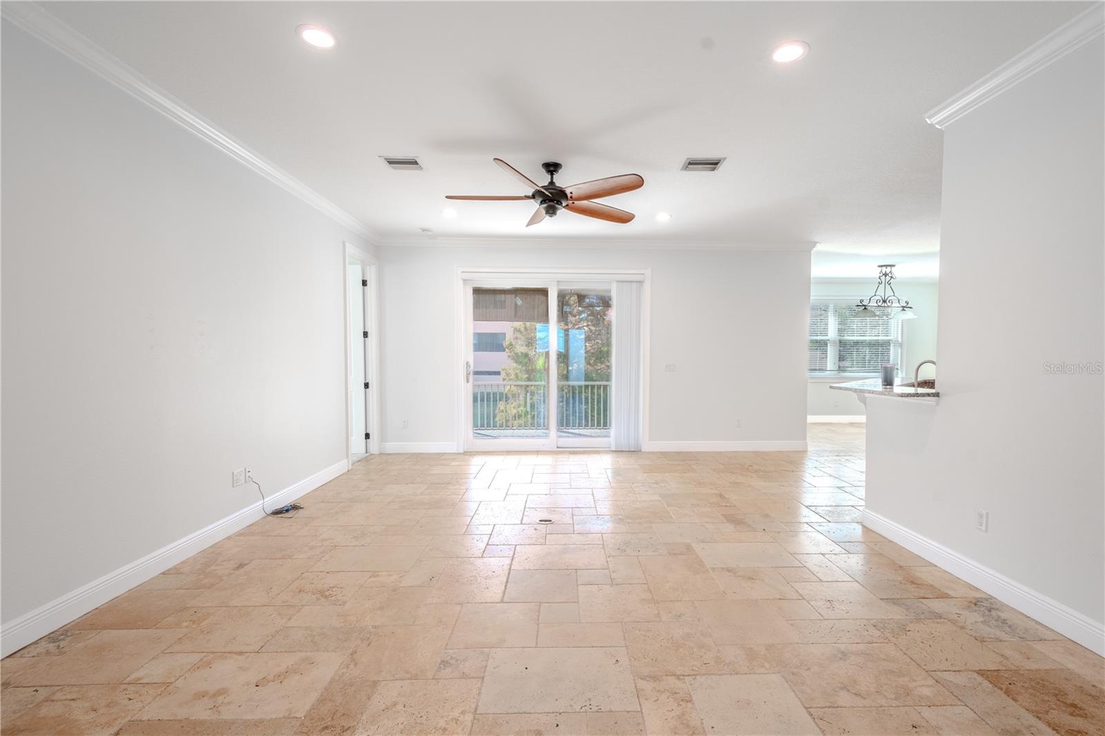 The expansive living room, adorned in soft neutral tones, showcases travertine flooring, recessed lighting, a ceiling fan, and sliding glass doors that open to the balcony, seamlessly blending indoor and outdoor living.