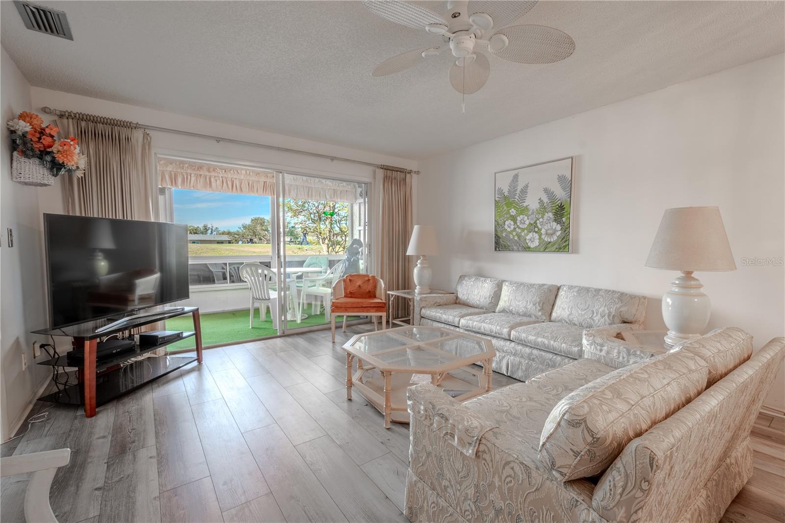 The living room features neutral tones, wood laminate flooring, a ceiling fan and sliding glass doors to the lanai wth a view of the golf course.