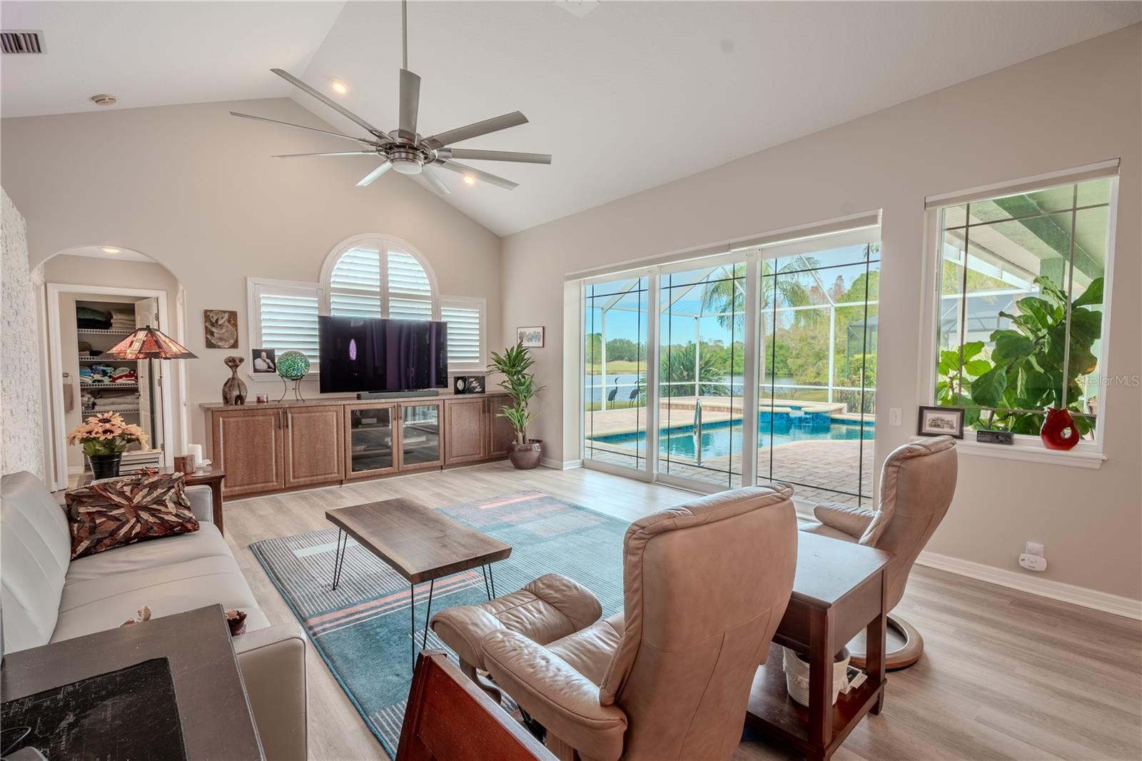 The family room features a cathedral ceiling with modern, sleek ceiling fan, luxury vinyl plank flooring, and views of the pool, pond and golf course.