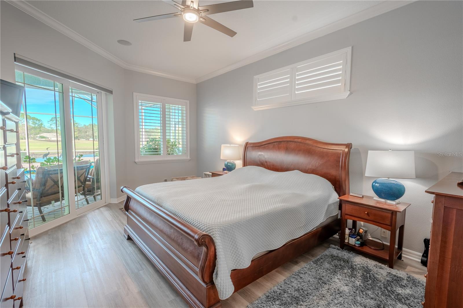 The primary bedroom is bathed in a neutral color palette, crown molding, and features a sleek ceiling fan with light kit, luxury vinyl flooring, an ensuite bath and glass doors opening to the lanai.