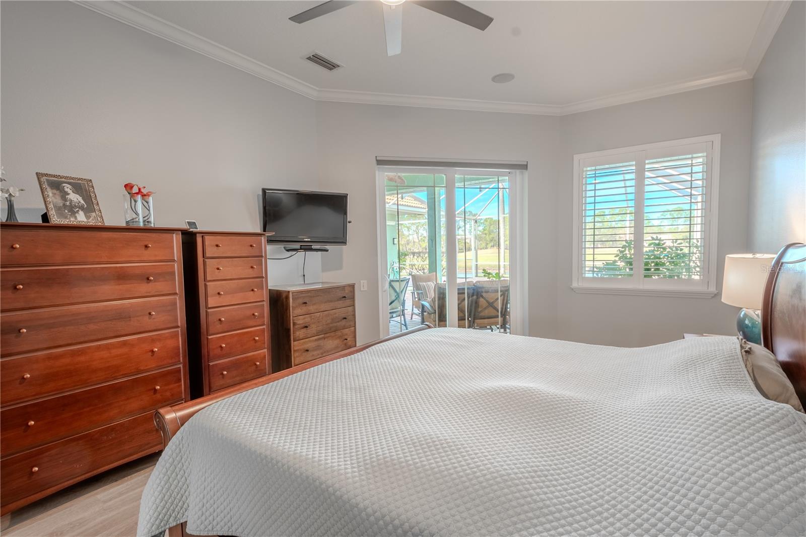 The primary bedroom is bathed in a neutral color palette, crown molding, and features a sleek ceiling fan with light kit, luxury vinyl flooring, an ensuite bath and glass doors opening to the lanai.