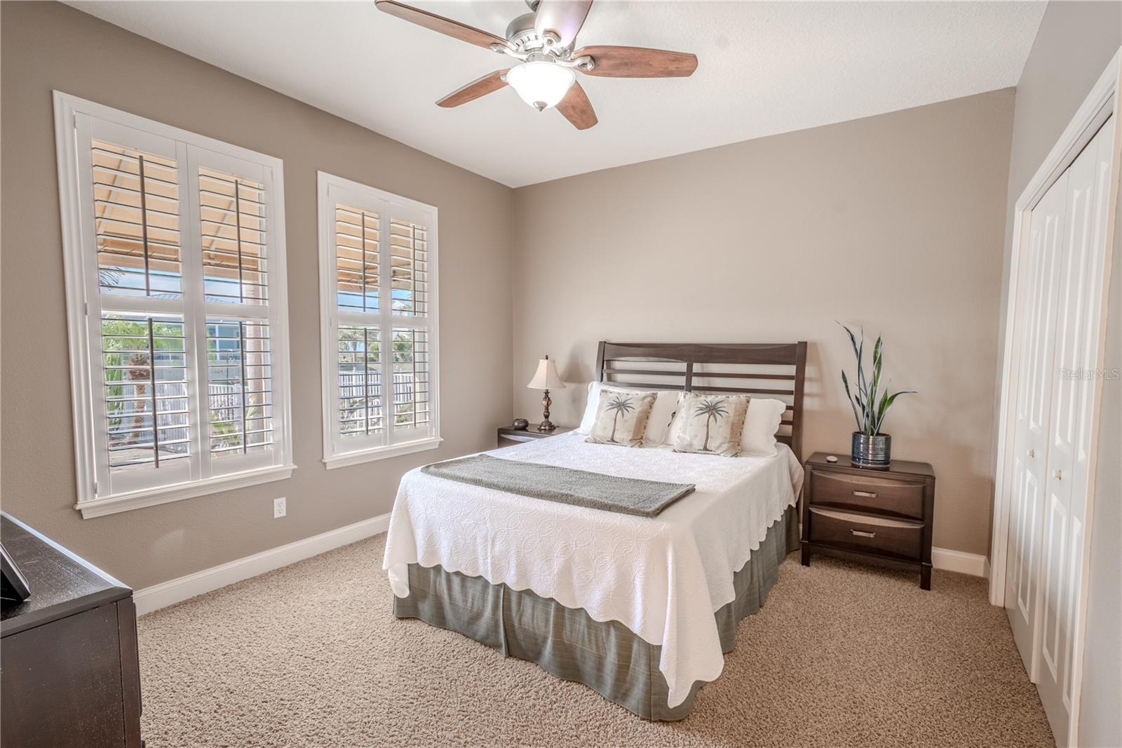 Bedroom 2 is on the opposite side of the primary bedroom on the first floor. It features warm tones, wood shutters, plush carpet, a ceiling fan and built in closet.