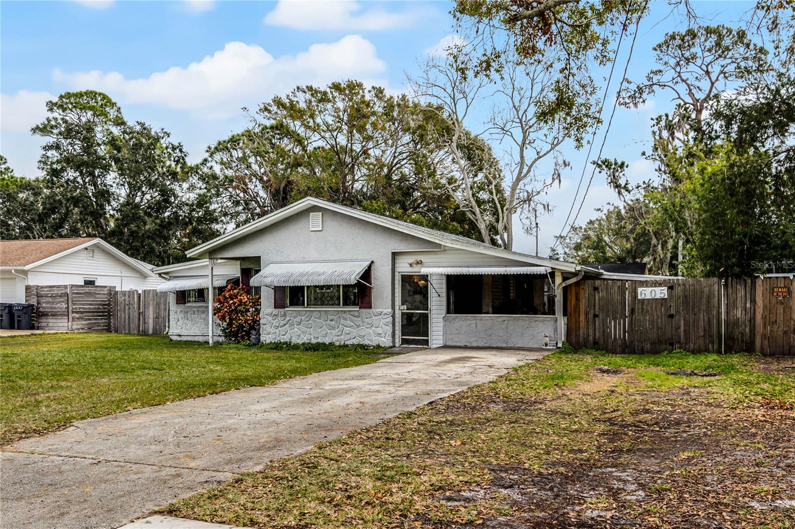 This charming single-story home features a light exterior with decorative stone accents and a gabled roof. The front elevation includes white awnings over the windows and a small covered porch area near the main entrance, adding character and shade. A paved driveway leads up to the right side of the home, while the left side features a well-maintained grassy yard bordered by a wooden fence. The home's classic design and quaint details create a welcoming curb appeal, ideal for those seeking a cozy and functional property.