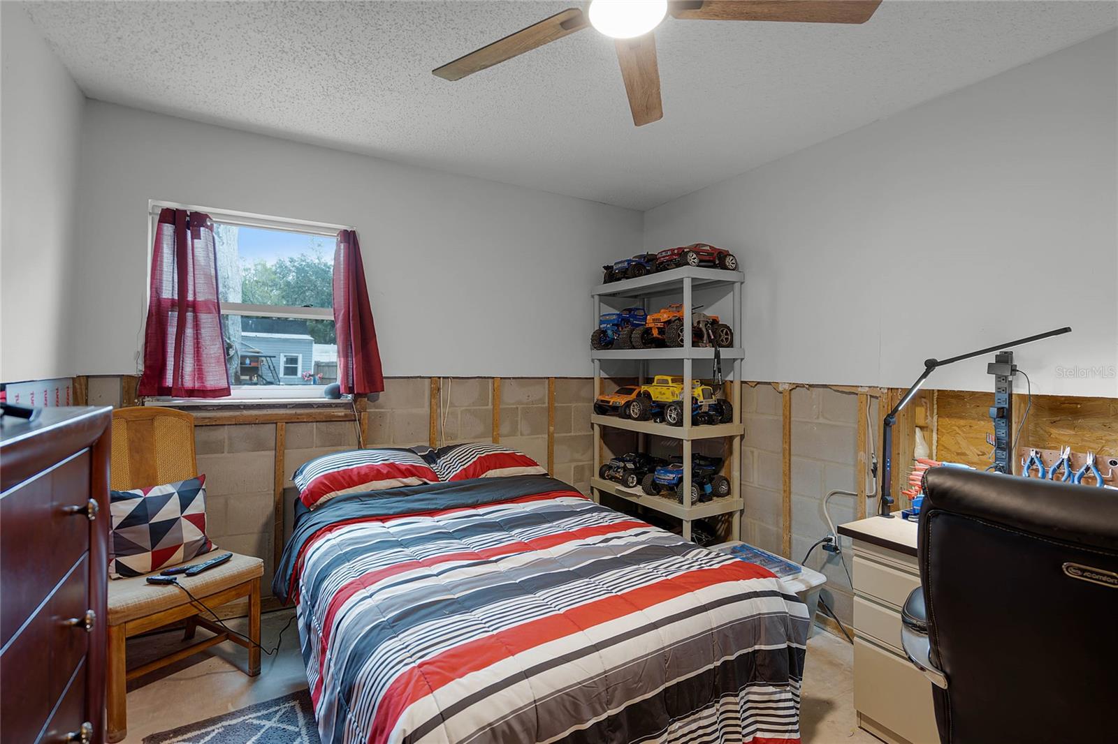 Bedroom with a bed featuring a striped red, black, and white comforter. A window with red curtains provides natural light, while a ceiling fan with a light fixture enhances the space. A shelving unit showcases a collection of model cars, and a desk with a task lamp is situated next to a black office chair. The lower portion of the walls has been stripped of drywall, approximately 4 feet from the floor, as part of flood remediation. The room has a concrete floor and a wooden chair with a decorative pillow.