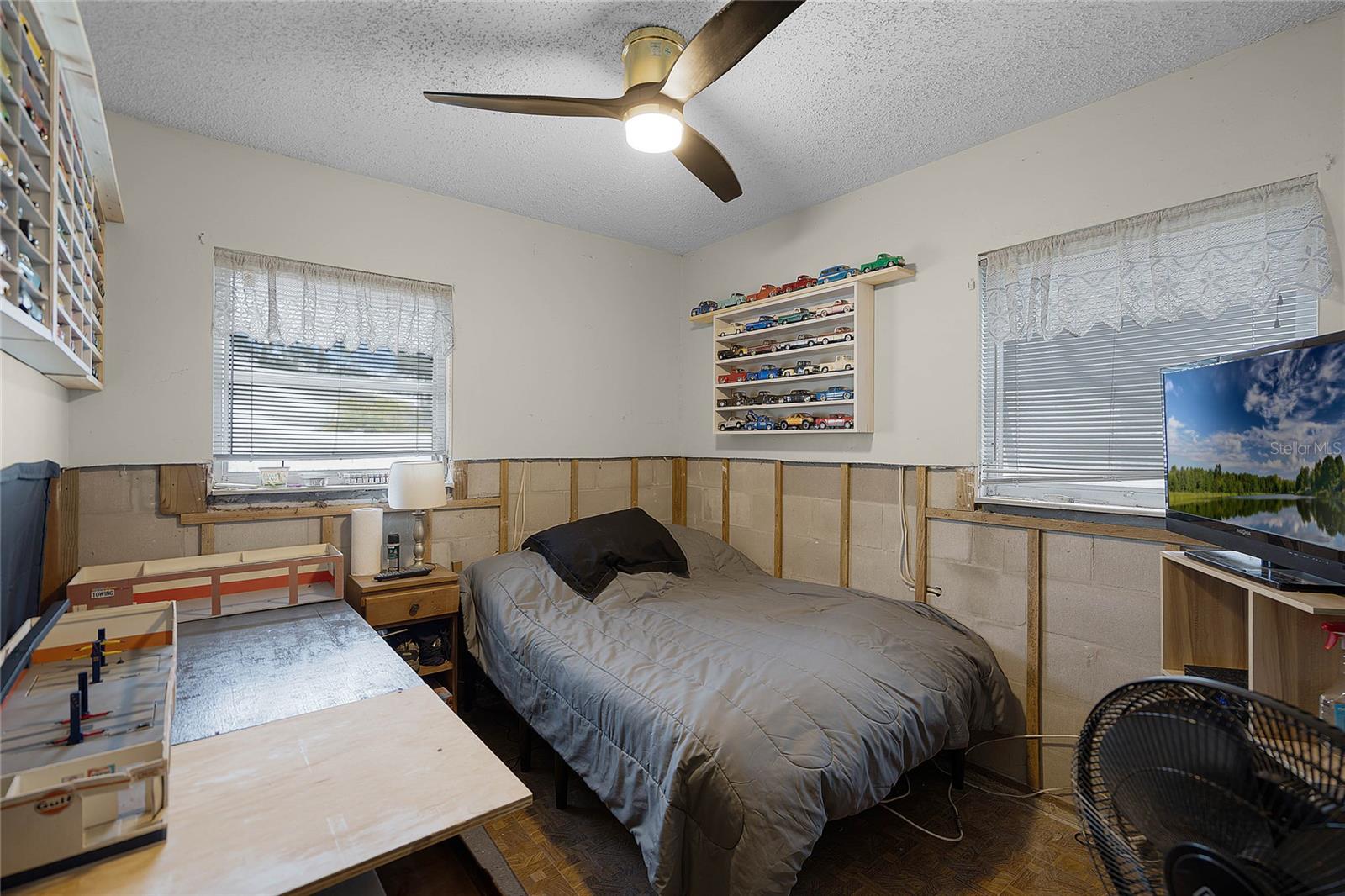 Cozy bedroom featuring a bed with a gray comforter and black pillow, positioned between two windows with lace curtains. A ceiling fan with a modern light fixture provides illumination. The walls display a collection of model cars on wooden shelves, while a large TV sits on a stand in the corner. A crafting or workspace table is set up next to the bed, with a small nightstand holding a lamp and other items. Partial drywall removal is visible along the lower walls, approximately 4 feet from the floor, as part of flood restoration efforts. The room has parquet flooring.