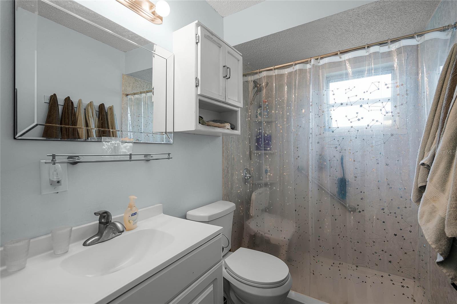 Bathroom with a single-sink vanity, a mirrored medicine cabinet, and additional storage above the toilet. The shower features a clear curtain with a constellation pattern and a window that allows natural light to brighten the space. A wall-mounted towel rack holds neatly arranged towels, and the room includes light blue walls and a standard toilet. The area has a clean and functional design.