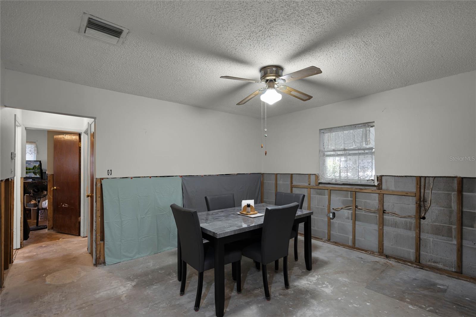 The entryway leads directly into the dining room, which features a central light fixture with a ceiling fan. The space includes a dining table with seating for four. Due to recent hurricane flooding, the walls have been partially stripped, with approximately 4 feet of drywall removed, revealing the cinder block structure and insulation. A small window brings natural light into the room, enhancing its potential for restoration and redesign.