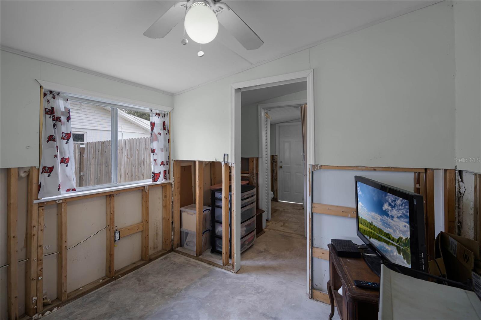 Third bedroom connecting to a half bath and utility room, featuring light green walls and a ceiling fan with a globe light. A large window with sports-themed curtains overlooks the fenced backyard, while partial drywall removal, approximately 4 feet from the floor, is visible along the lower walls due to flood remediation. The room has a concrete floor and contains a small table with a television and shelving for storage. The layout offers functional access to adjacent spaces.