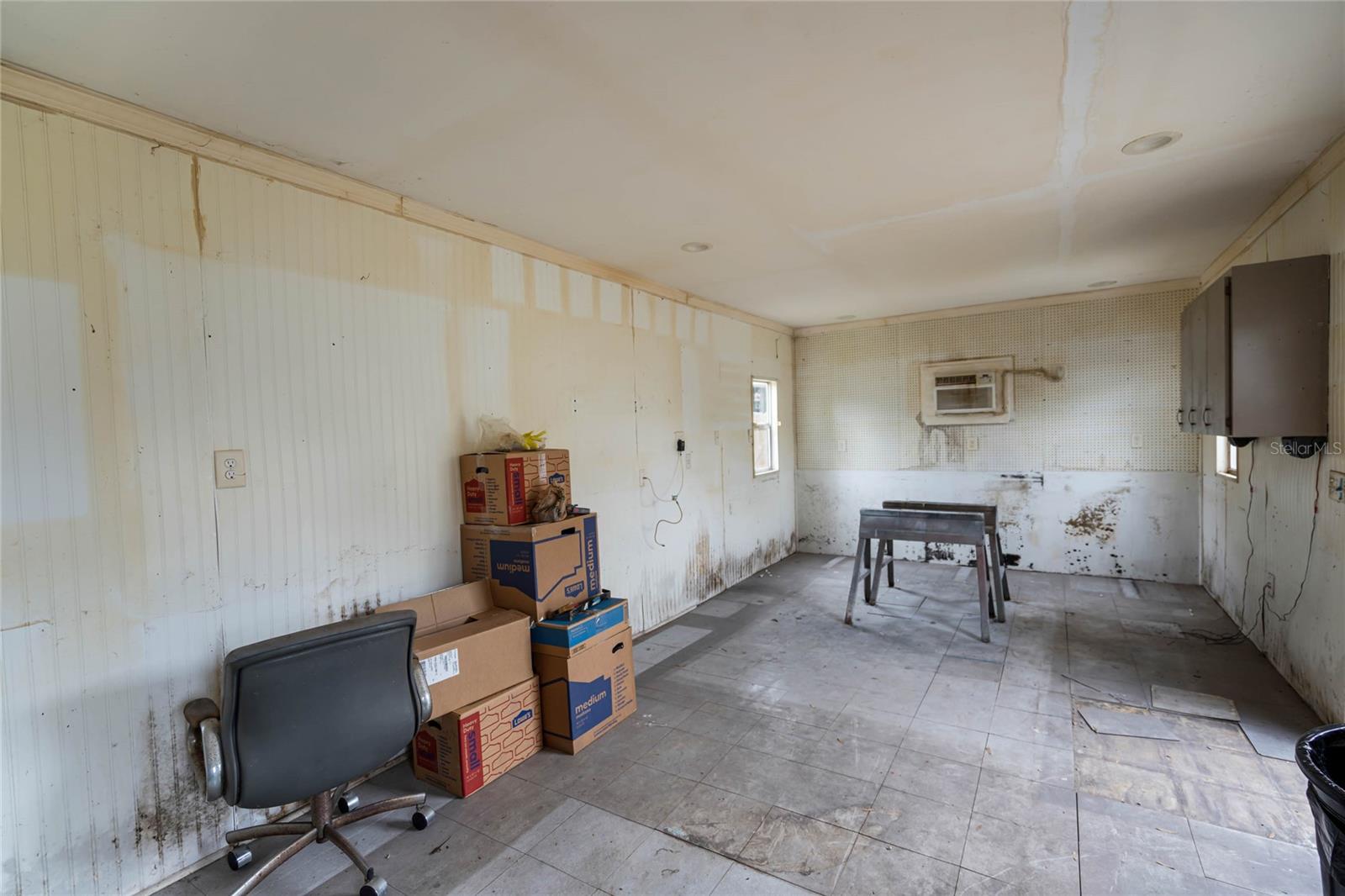 Attached workshop or storage area with white paneled walls and visible signs of water damage near the lower sections. The space features tile flooring and built-in cabinets along one wall, with a small window and wall-mounted air conditioning unit. A stack of moving boxes and a rolling office chair are placed along one side, and a work table sits in the center of the room. The area offers versatile utility but will require restoration to address existing damage.