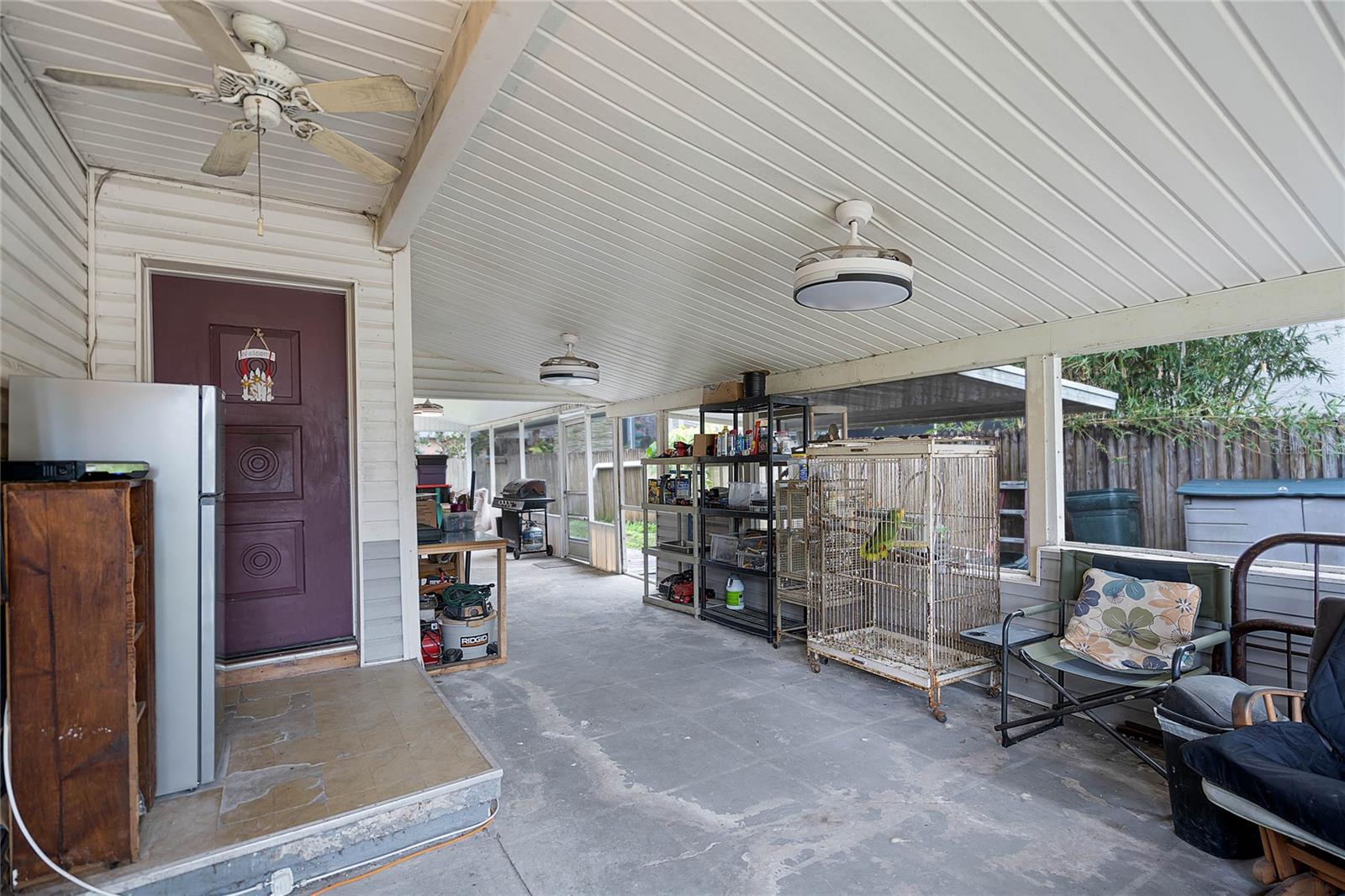 Covered patio with a white paneled ceiling and multiple ceiling fans for ventilation. The space features a purple door leading to the interior, shelving units for storage, and a large birdcage. A mix of outdoor furniture, including chairs with floral cushions, provides seating. The patio offers ample space for activities and includes a view of the fenced backyard. Concrete flooring throughout shows signs of wear, and a grill is situated at one end of the area for outdoor cooking.