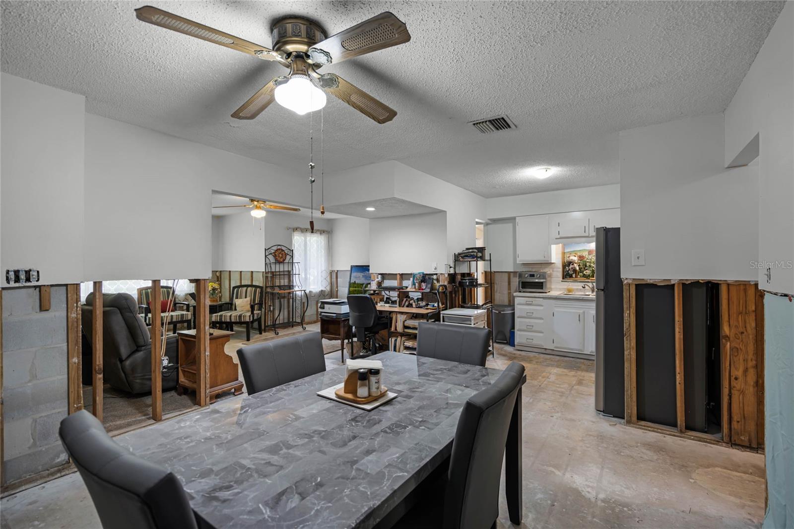 The dining area showcasing partially stripped drywall approximately 4 feet from the floor due to recent flood remediation. The room features a dining table with a marble-patterned surface, gray upholstered chairs, and a ceiling fan with a light. The adjacent kitchen includes white cabinetry and modern appliances. The space transitions into a seating area with natural light streaming through sheer curtains, highlighting the home's potential after necessary restoration.