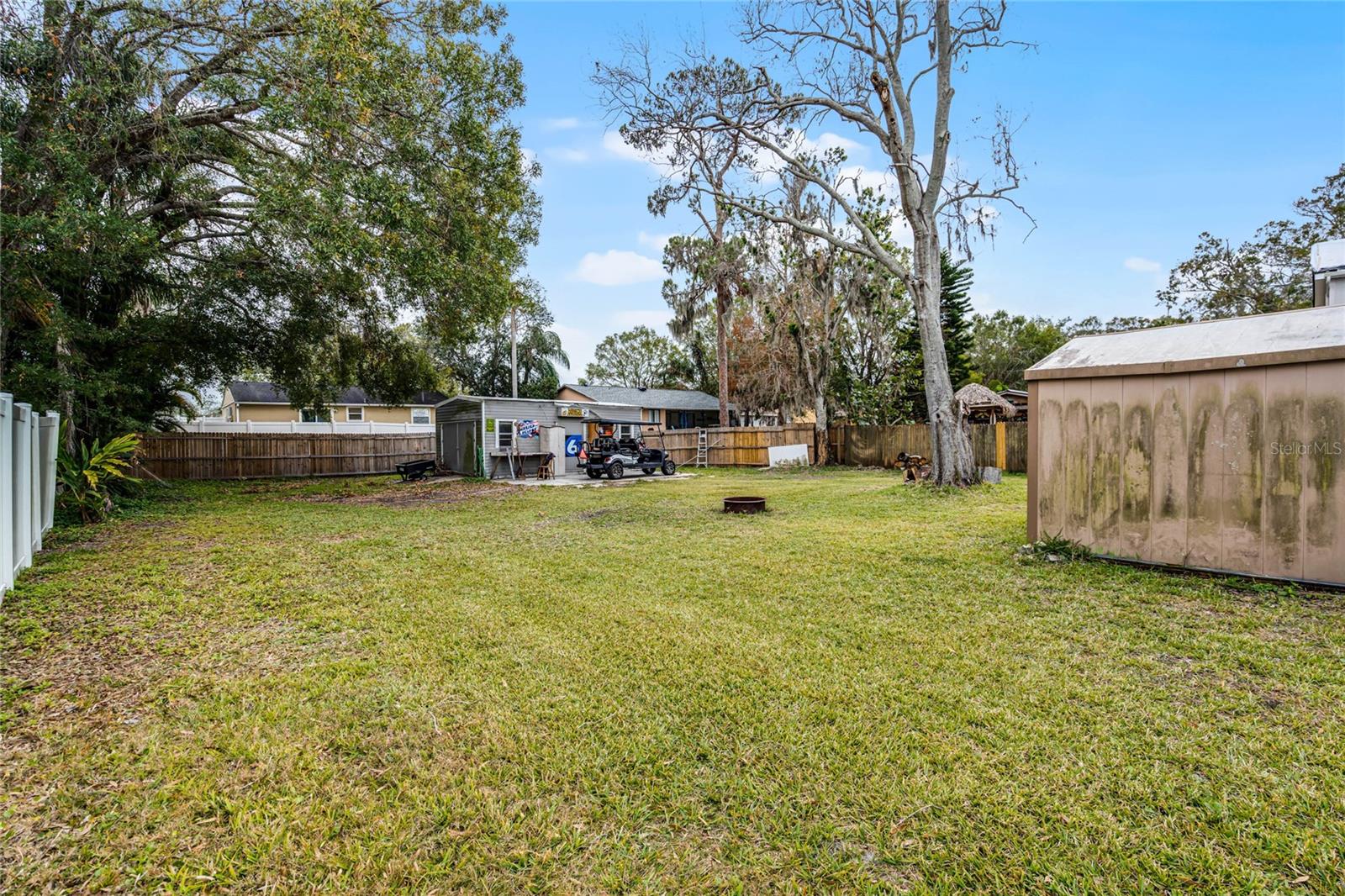 Spacious backyard with a large grassy area surrounded by a wooden privacy fence. The yard includes a mature tree providing shade, a fire pit, and two storage structures—one metal shed and one wooden outbuilding. A paved area with a golf cart and additional storage space is located near the back of the property. The yard offers ample space for outdoor activities and potential for customization.