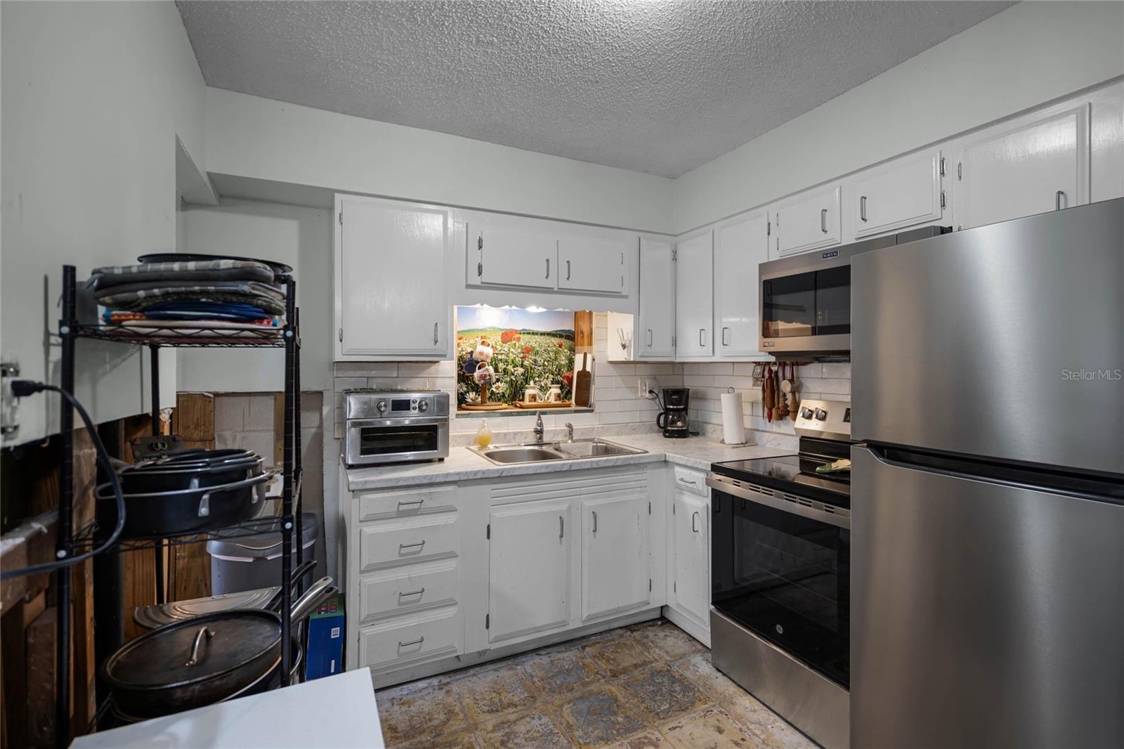 Bright kitchen featuring white cabinetry, a double stainless-steel sink, and modern appliances, including a stainless-steel refrigerator, microwave, and oven. A backsplash of white subway tile complements the countertop, and a large window above the sink showcases a colorful outdoor view. A metal shelving unit adds additional storage, with partial drywall removal visible along the lower walls as part of flood restoration efforts. Tile flooring throughout the space.