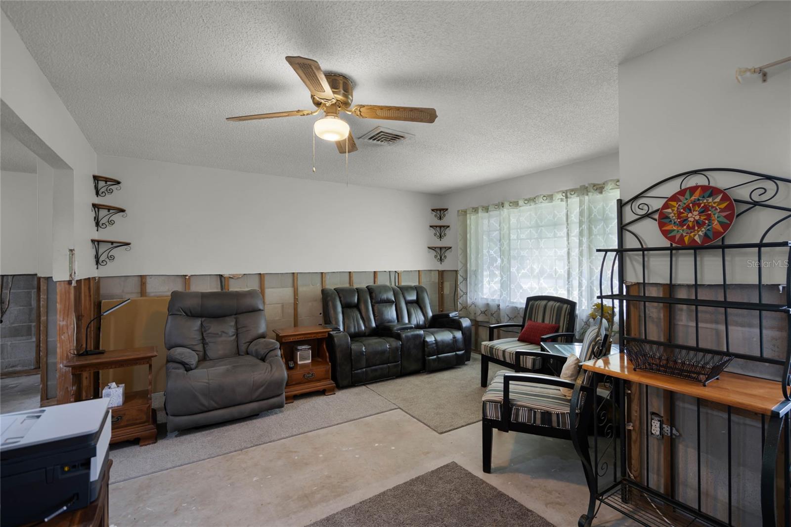 Living room with a ceiling fan and large windows framed by sheer patterned curtains, allowing natural light to brighten the space. Furnished with a gray recliner, black leather sofa, and striped armchairs accented by red pillows. A decorative metal shelving unit adds character to the room. Partial drywall removal, approximately 4 feet from the floor, is visible due to flood remediation, with exposed concrete and wood along the lower walls. The room features a mix of area rugs and concrete flooring.