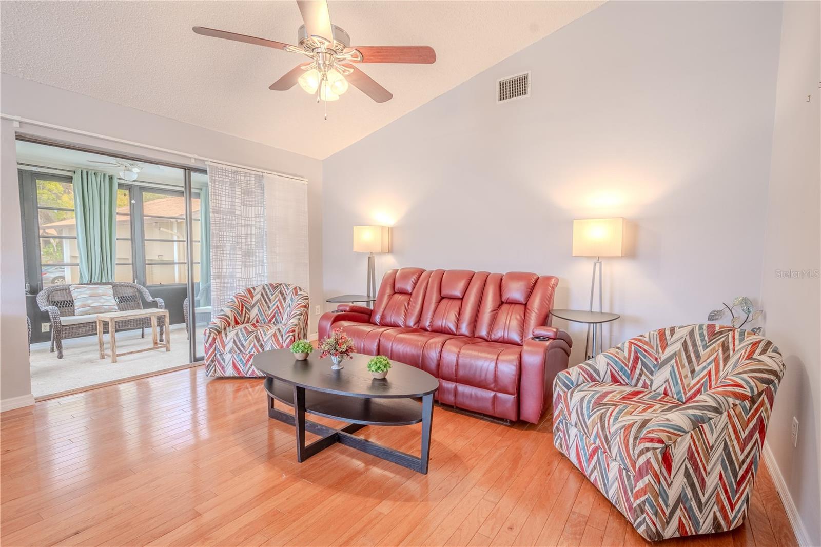 The living room features wood flooring,a vaulted ceiling with ceiling fans, neutral tones and sliding glass doors to the enclose lanai/Florida Room.