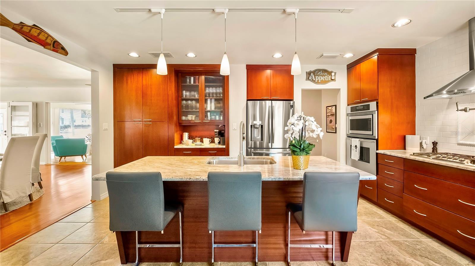 Stunning modern kitchen featuring rich wood cabinetry, sleek stainless steel appliances, a spacious center island with seating, and elegant pendant lighting, seamlessly flowing into the bright and airy living spaces.