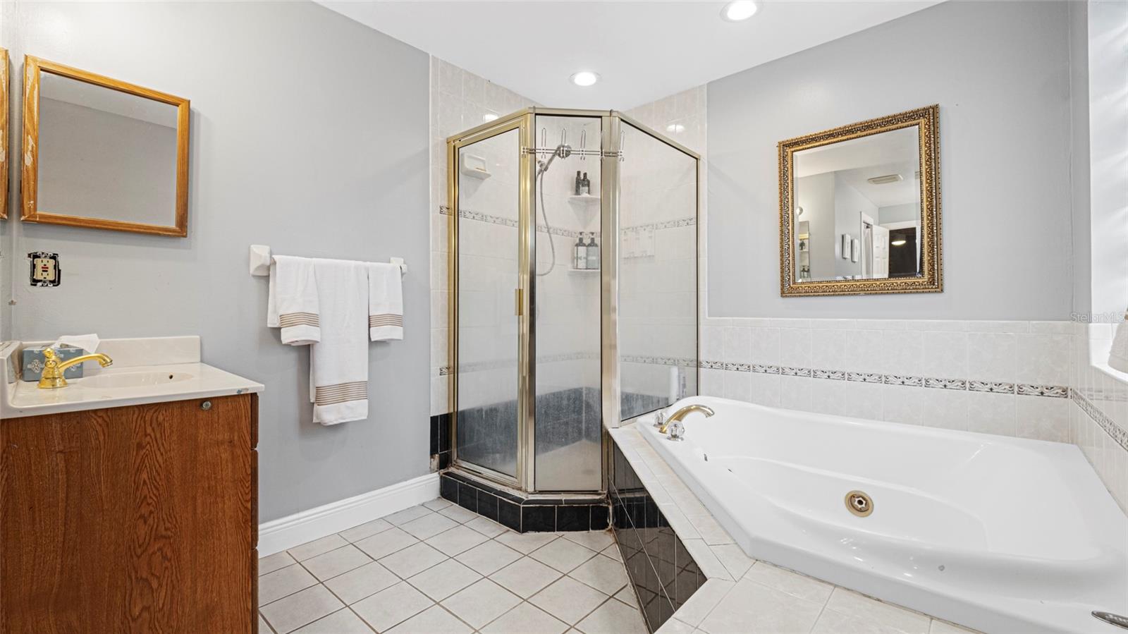 Spacious primary bathroom that offers a blend of comfort and functionality, featuring a large jetted soaking tub, a separate glass-enclosed shower with gold-tone trim and dual vanities.