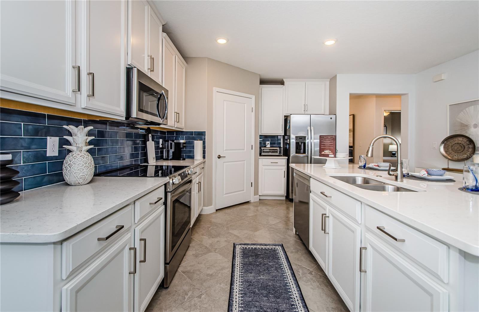 The striking blue tile backsplash adds a touch of sophistication, beautifully complementing the crisp white cabinetry with modern brushed hardware. Thoughtfully designed with ample storage, a spacious pantry, and a large center island, this kitchen is perfect for both casual meals and entertaining. The open flow leads seamlessly into the dining and living areas, making it the true heart of the home.