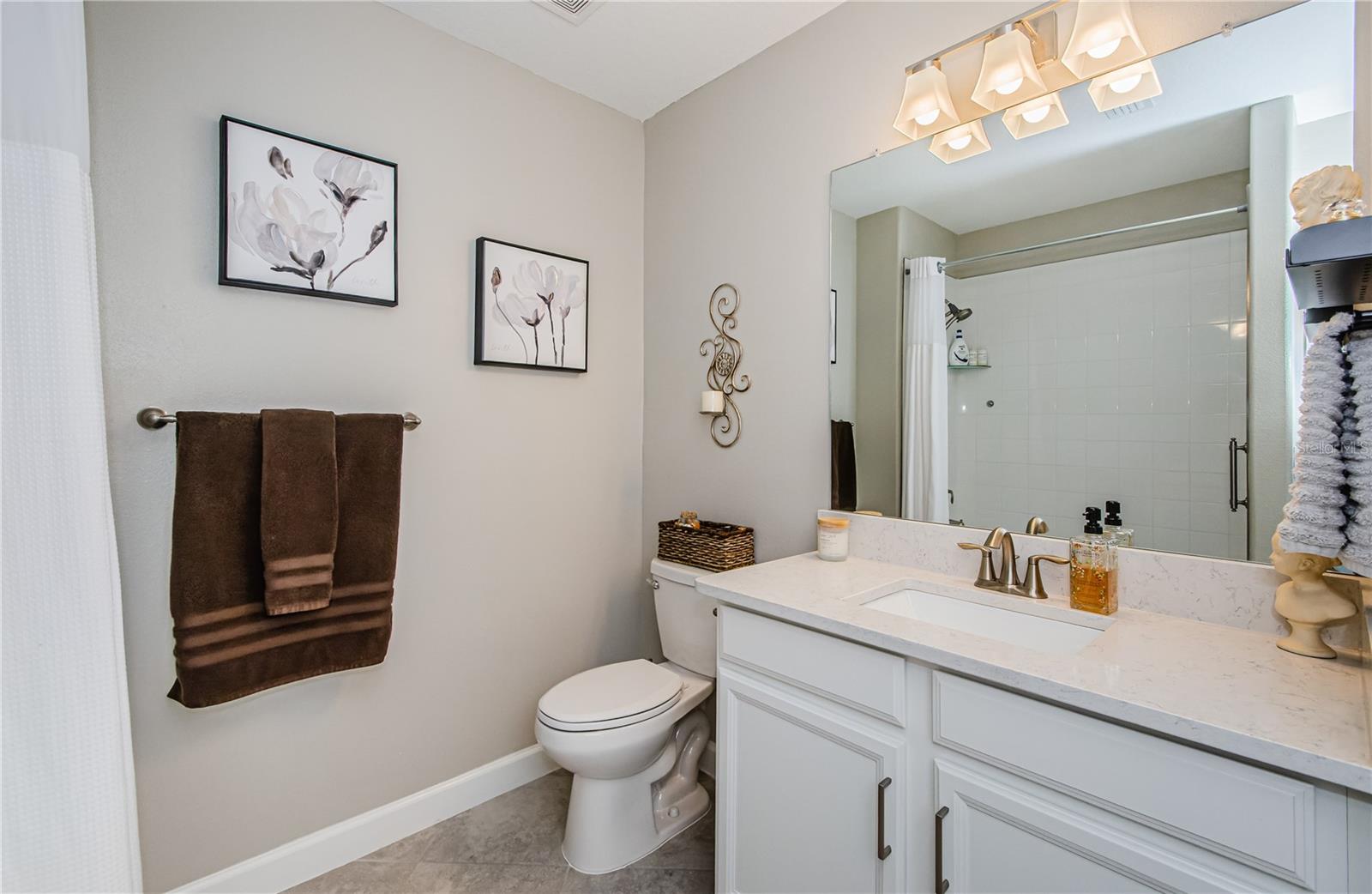 A spa-like oasis of relaxation, this beautifully designed bathroom offers an inviting neutral color palette that exudes warmth and sophistication. The quartz vanity with modern bronze fixtures, elegant framed floral artwork, and plush linens create an environment that’s both functional and stylish.