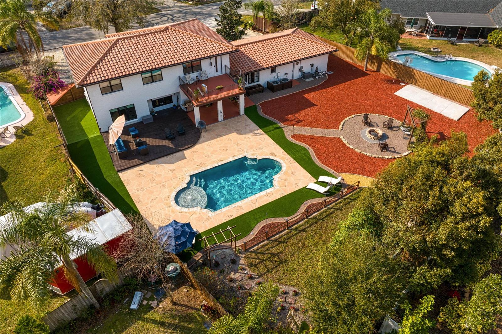 Aerial back yard exterior with Grecian style pool  custom lights, a curated fire pit lounge, wooden deck, and an expansive patio overlooking a whimsical butterfly garden.