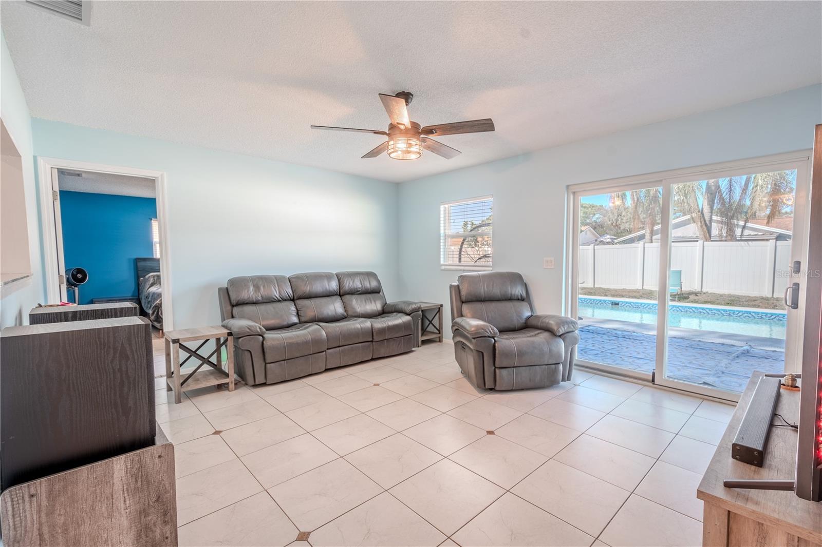 The living room features neutral tone ceramic tile floor, a ceiling fan with light kit, and sliding glass doors frame the view of the saltwater pool.