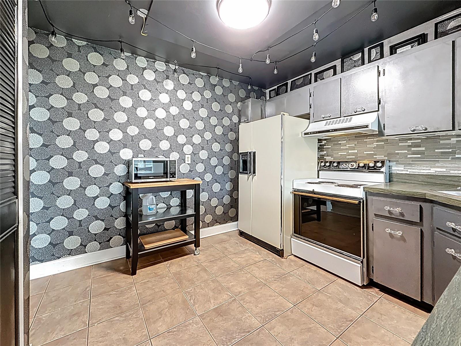 At the side of the breakfast bar, looking into the kitchen. The wall pantry is behind you, to the left.