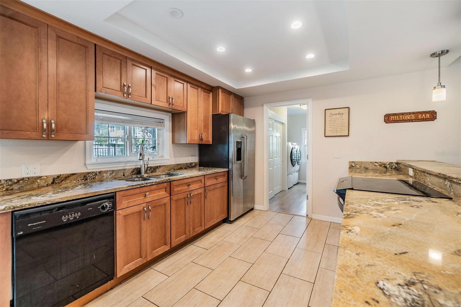 Beautifully updated kitchen with granite countertops, striking cherry wood cabinets, stainless steel stove and refrigerator, plank tile flooring, and elevated breakfast counter overlooking the living room.