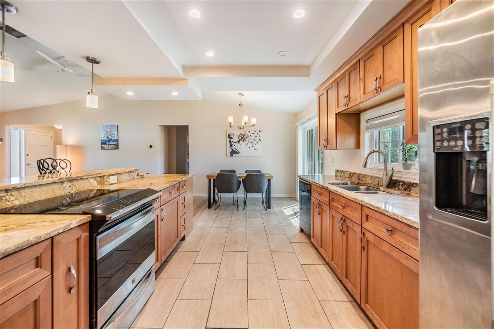 Beautifully updated kitchen with granite countertops, striking cherry wood cabinets, stainless steel stove and refrigerator, plank tile flooring, and elevated breakfast counter overlooking the living room.