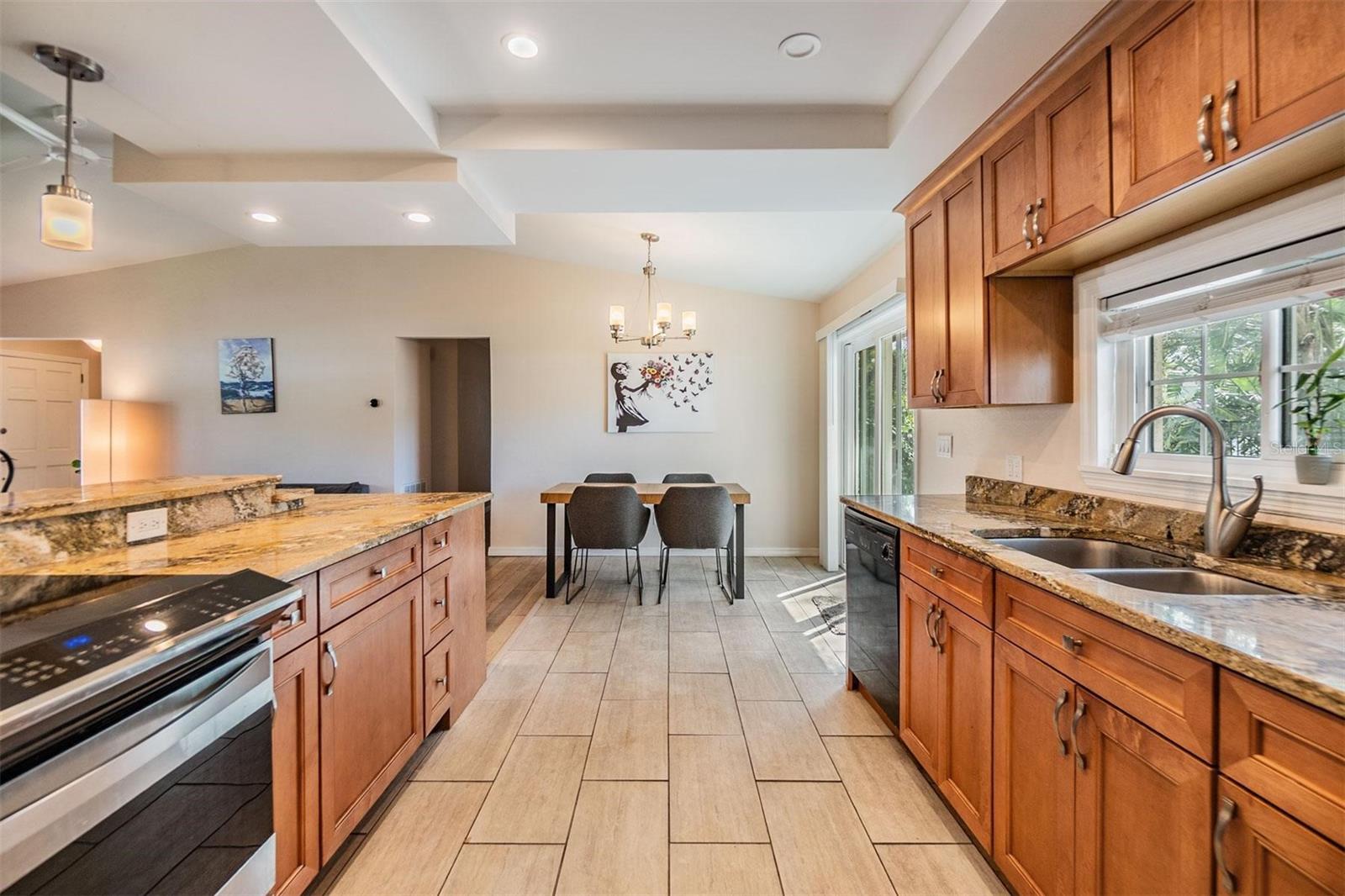 Beautifully updated kitchen with granite countertops, striking cherry wood cabinets, stainless steel stove and refrigerator, plank tile flooring, and elevated breakfast counter overlooking the living room.