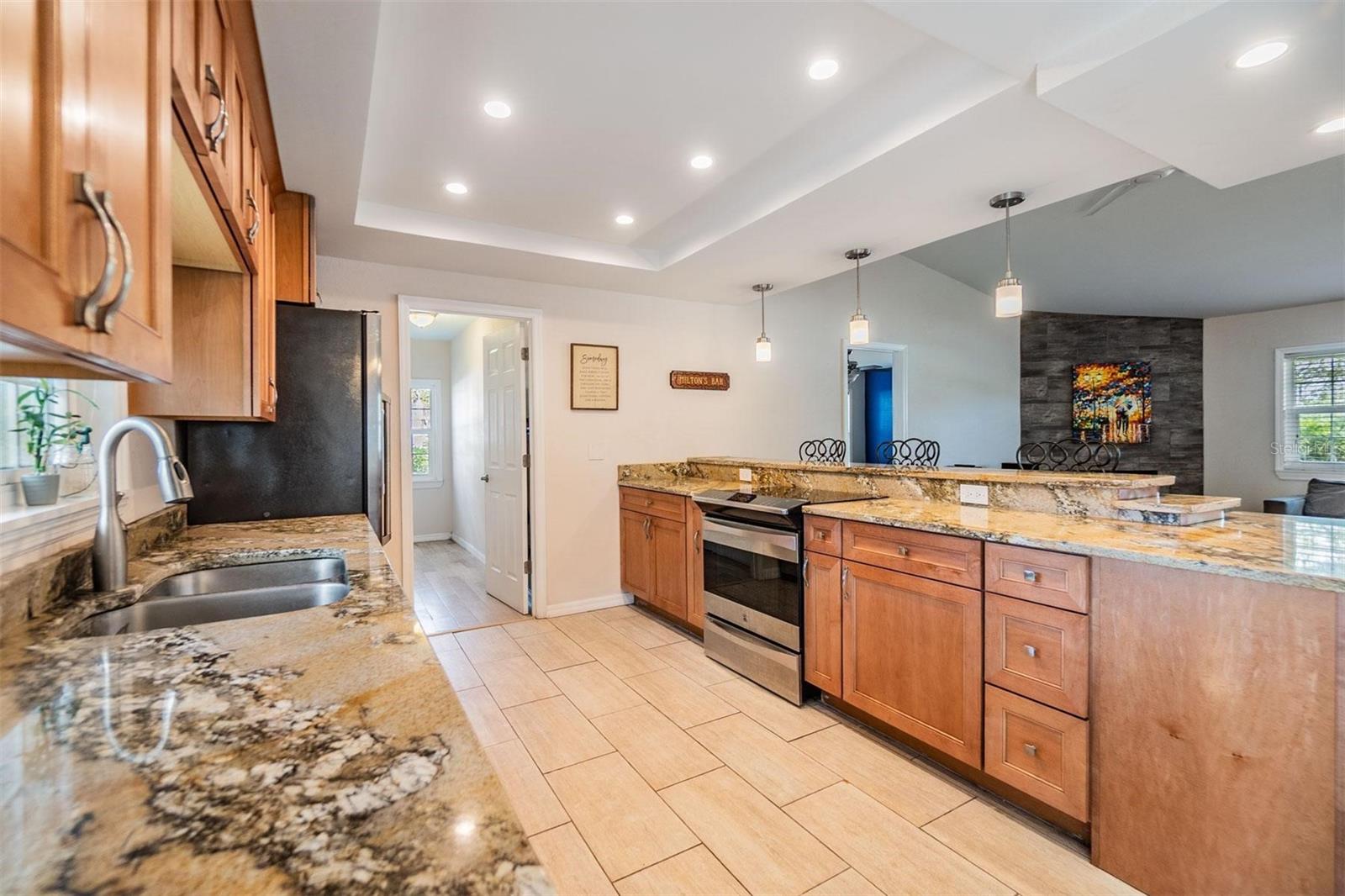 Beautifully updated kitchen with granite countertops, striking cherry wood cabinets, stainless steel stove and refrigerator, plank tile flooring, and elevated breakfast counter overlooking the living room.