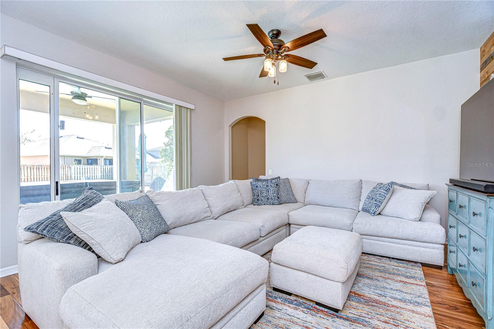 This living room radiates warmth with its inviting sun-drenched ambiance, thanks to expansive glass doors leading to the outdoor space