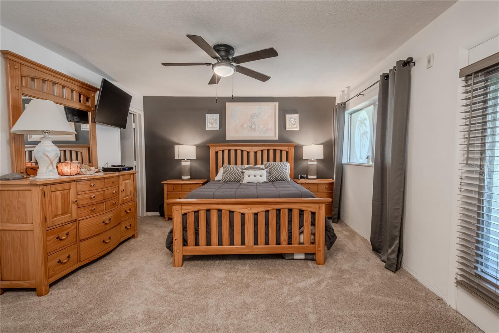 The bedroom features new carpet, neutral tones with an accent wall, rods and drapes, a walk-in closet, a modern ceiling fan with light kit, bed, nightstand, lamps, dresser and door to the lanai.