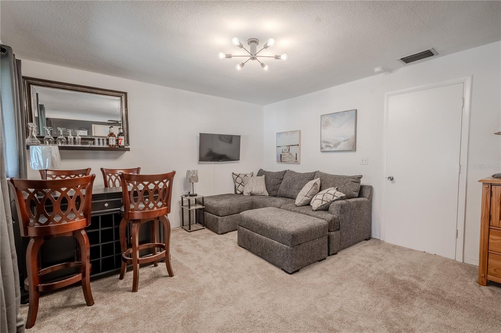 The bedroom features a Counter Height Console Dining Table with seating for 4, a new stylish lighting fixture, a love seat sectional with ottoman and a wall mount flat screen TV.