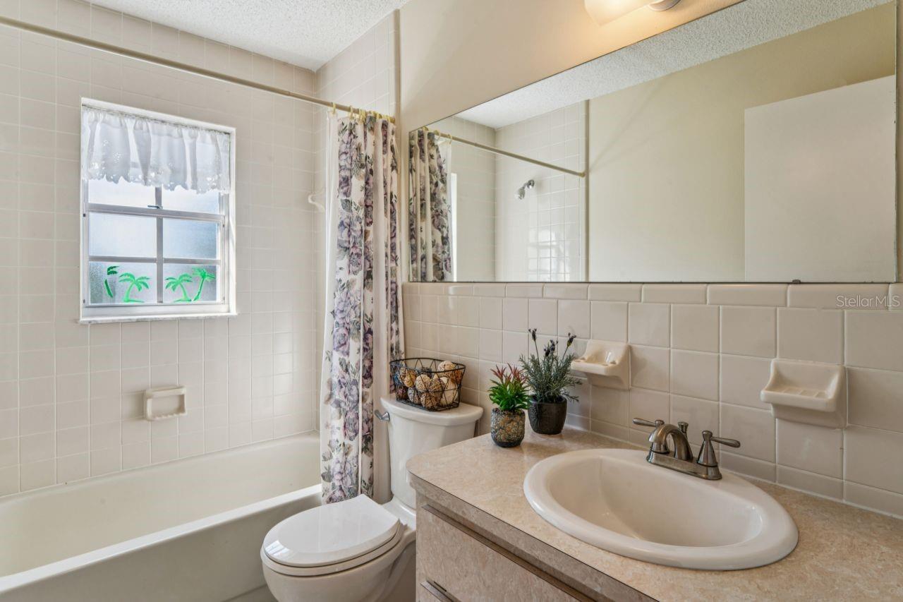 The guest bathroom features a single vanity, a commode, a bright window, a wall mirror, a stainless-steel faucet, and a full-sized bathtub.