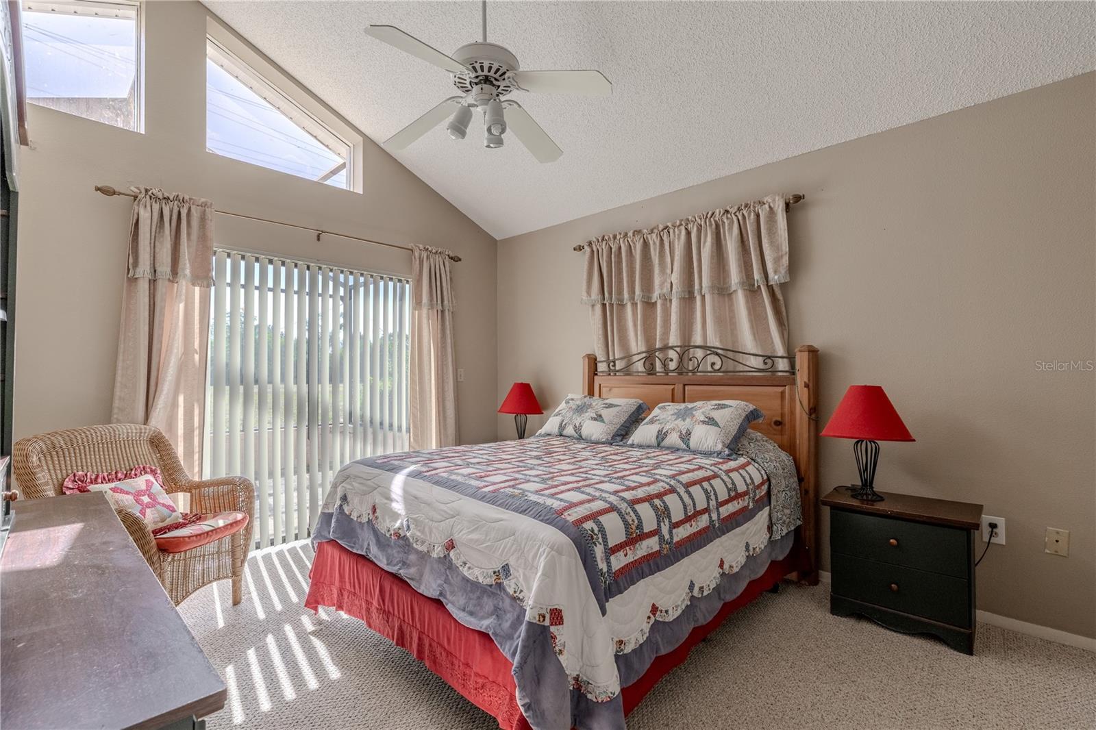 The primary bedroom features a vaulted ceiling, neutral tone walls and carper, a ceiling fan with light kit, and transom windows above a the sliding glass doors.